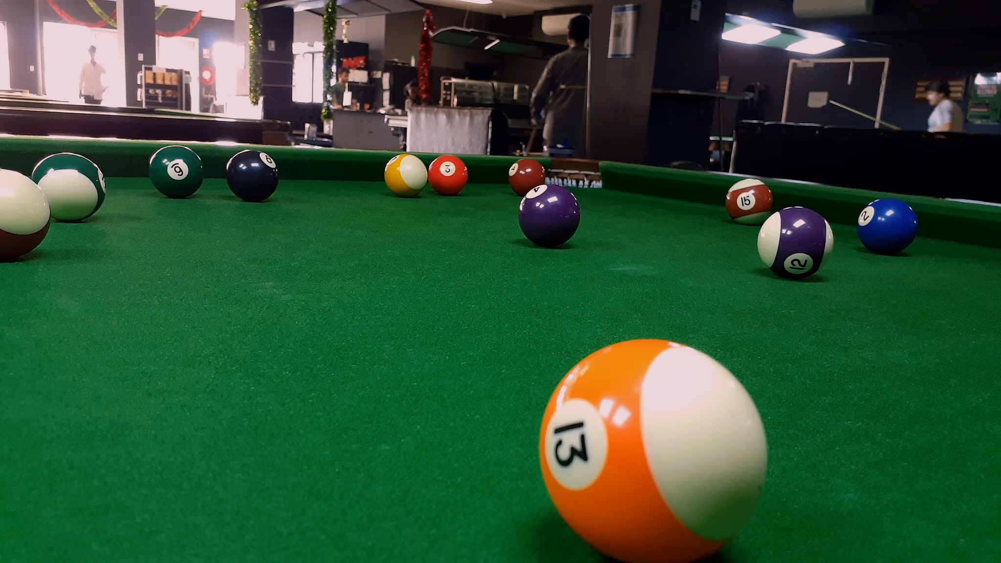 American pool balls scattered across green cloth in close focus, with the lounge's service counter and display cabinet out of focus beyond.