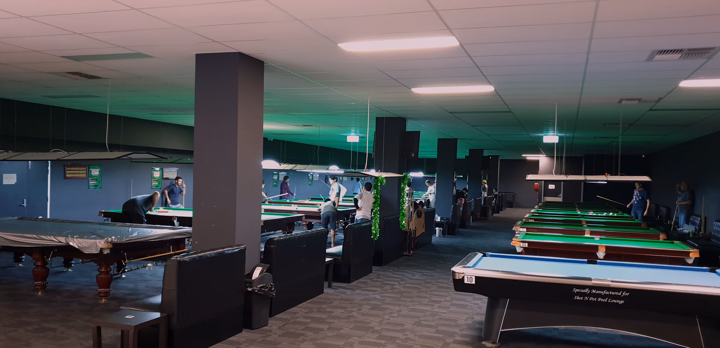 The American pool table on blue cloth, its rail lettered "Specially Manufactured for Shot N Pot Pool Lounge", with charcoal columns and the snooker tables beyond.