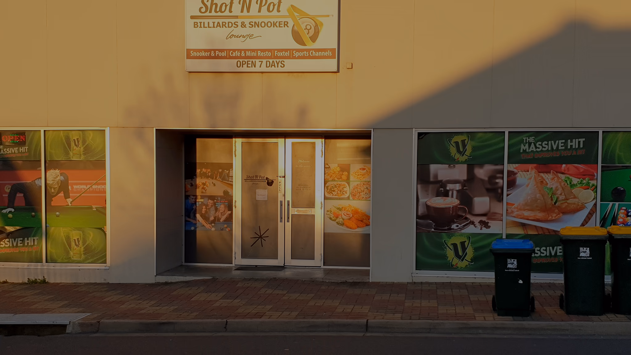 The glass entrance door under the Shot N Pot sign, poster windows either side, late sun across the wall.