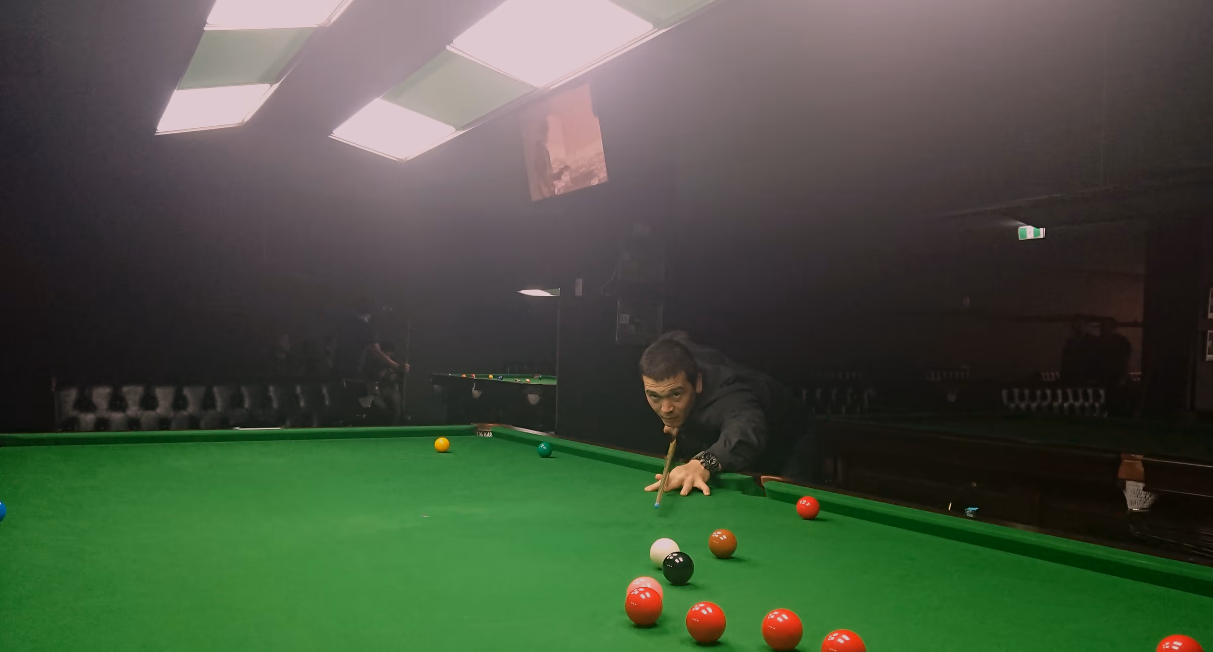 A single player at the far end of a snooker table, lit from directly above by the raft, the rest of the room dark around the pool of light.