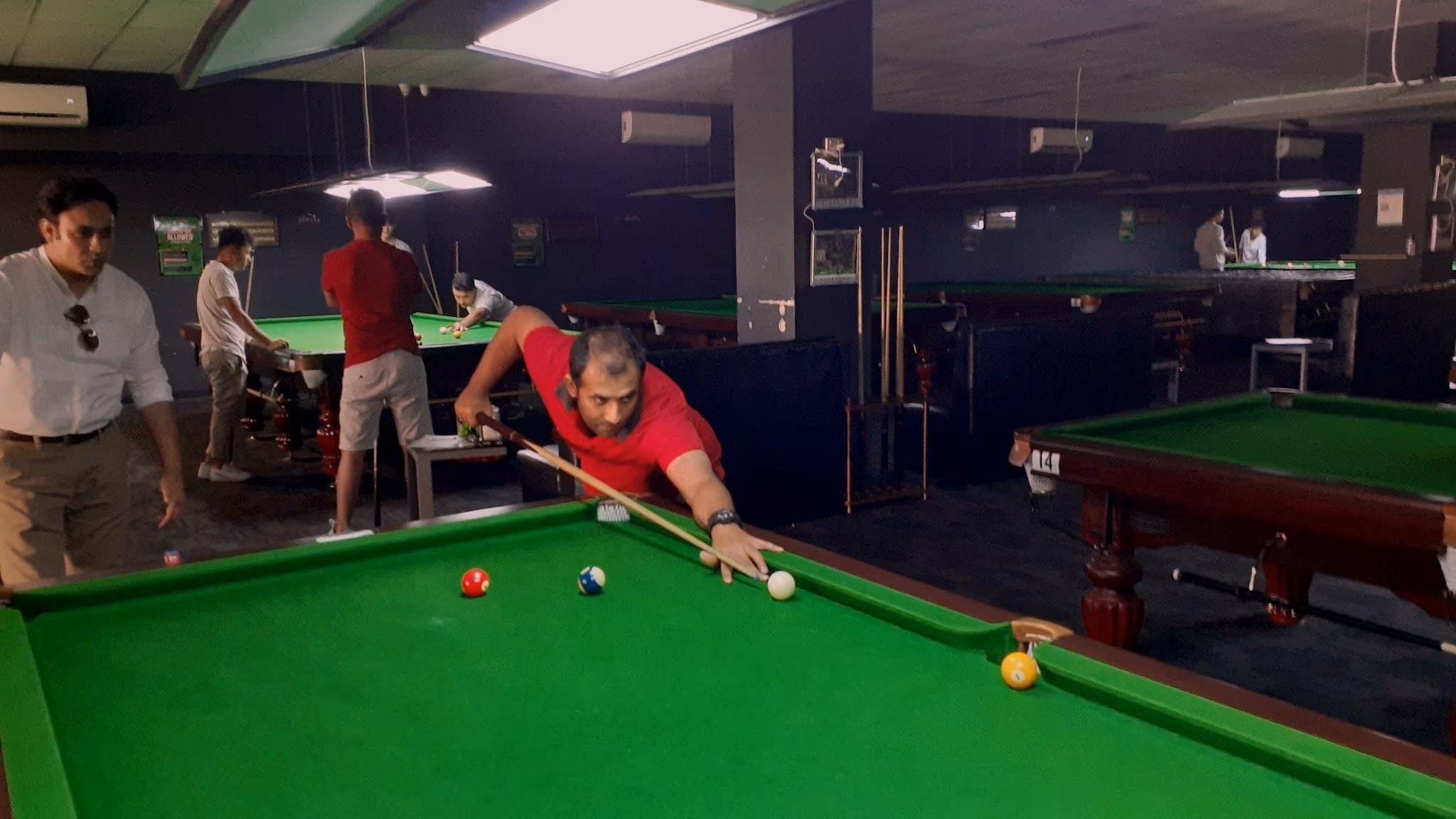 A busy snooker room. A player in a red shirt leans low over a shot on a full-size table in the foreground while others play at tables receding into the room behind him.
