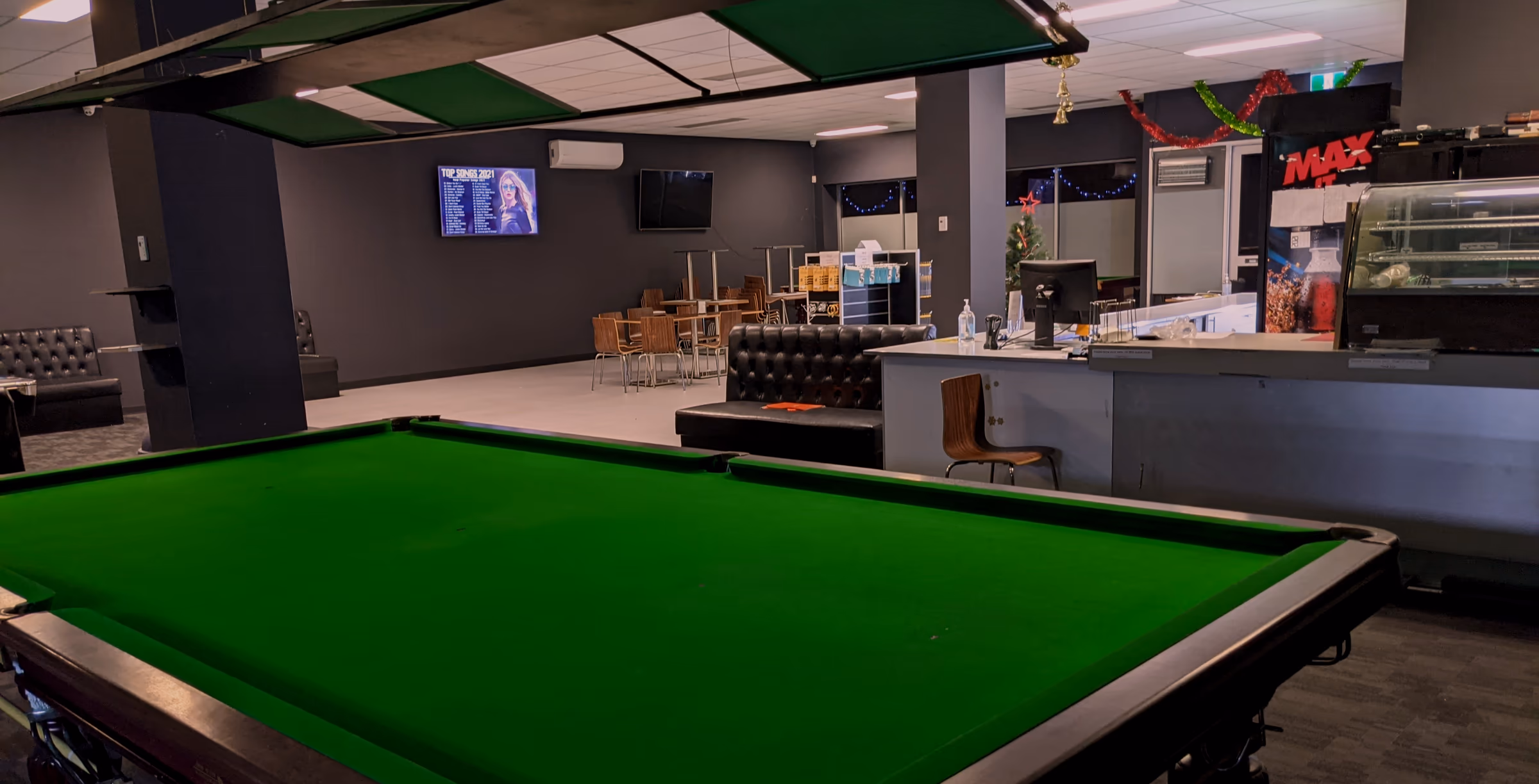 An empty snooker table in daylight with the lamps off, the green-topped light rafts hanging above it on their chains, a display cabinet and leather bench beyond.