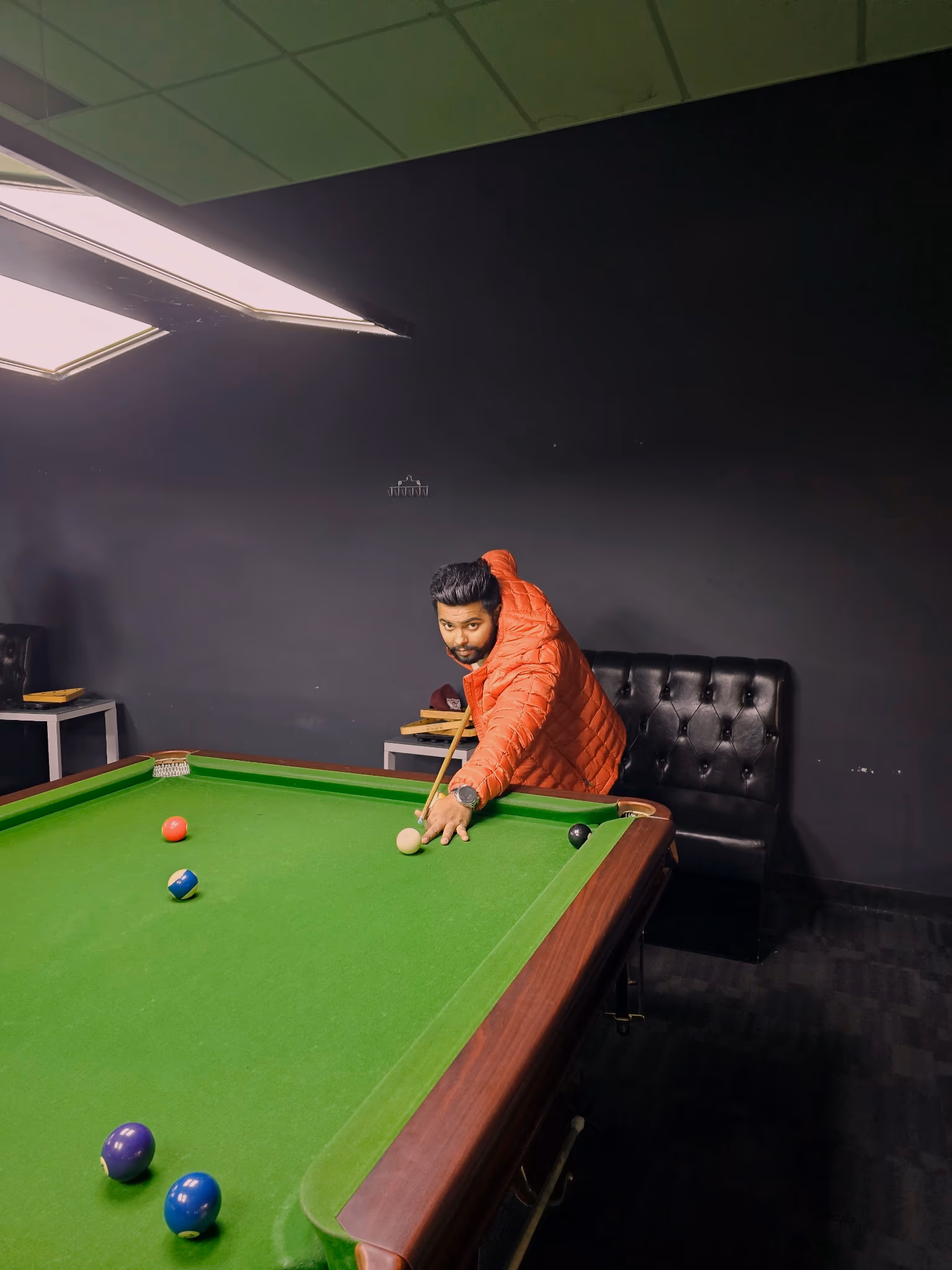 A player in an orange jacket lining up a shot at a snooker table, with a buttoned black leather bench seat against the wall behind him.