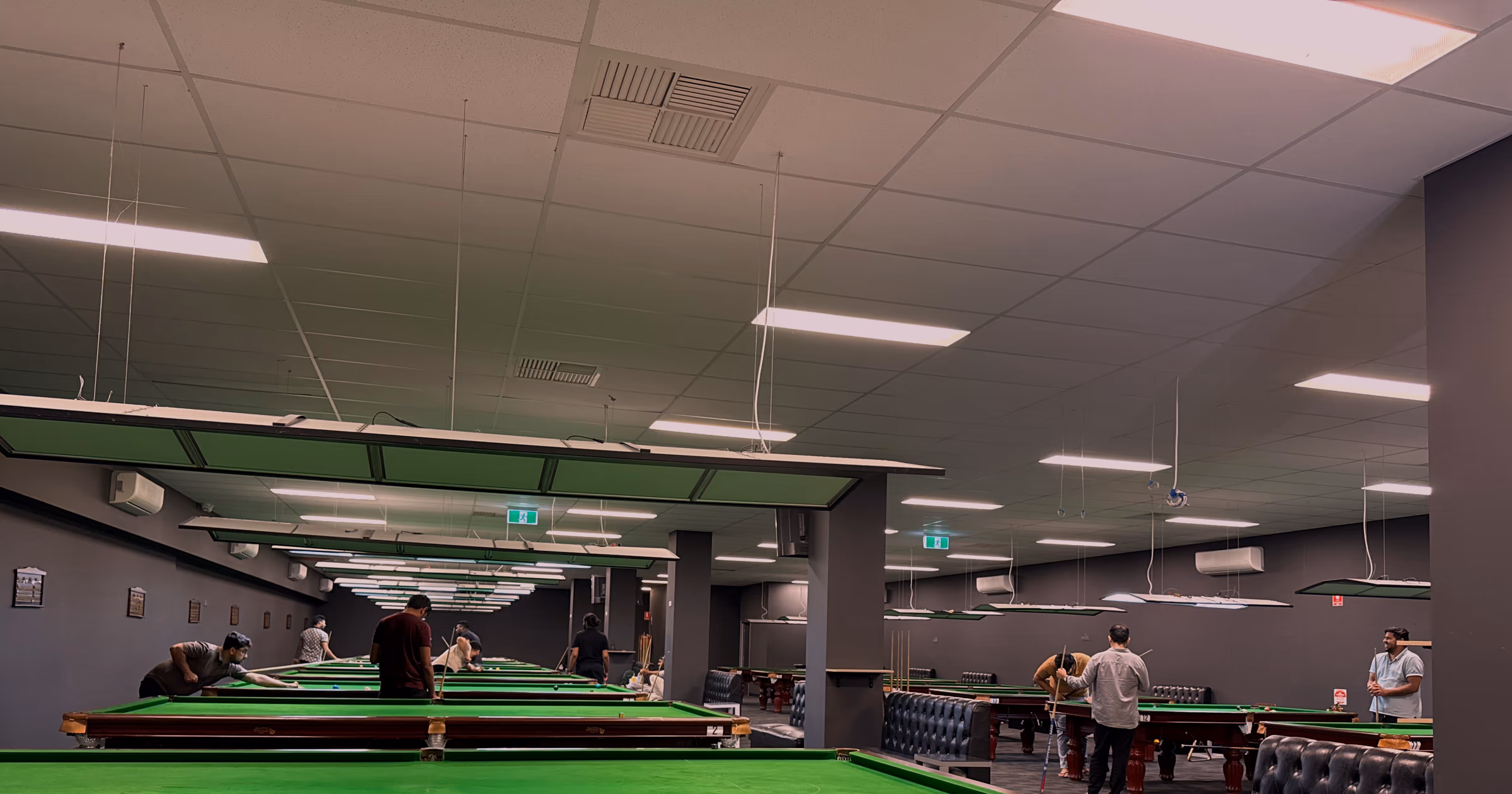 The row of suspended light rafts receding down the room above the tables, players small and far away at the end.