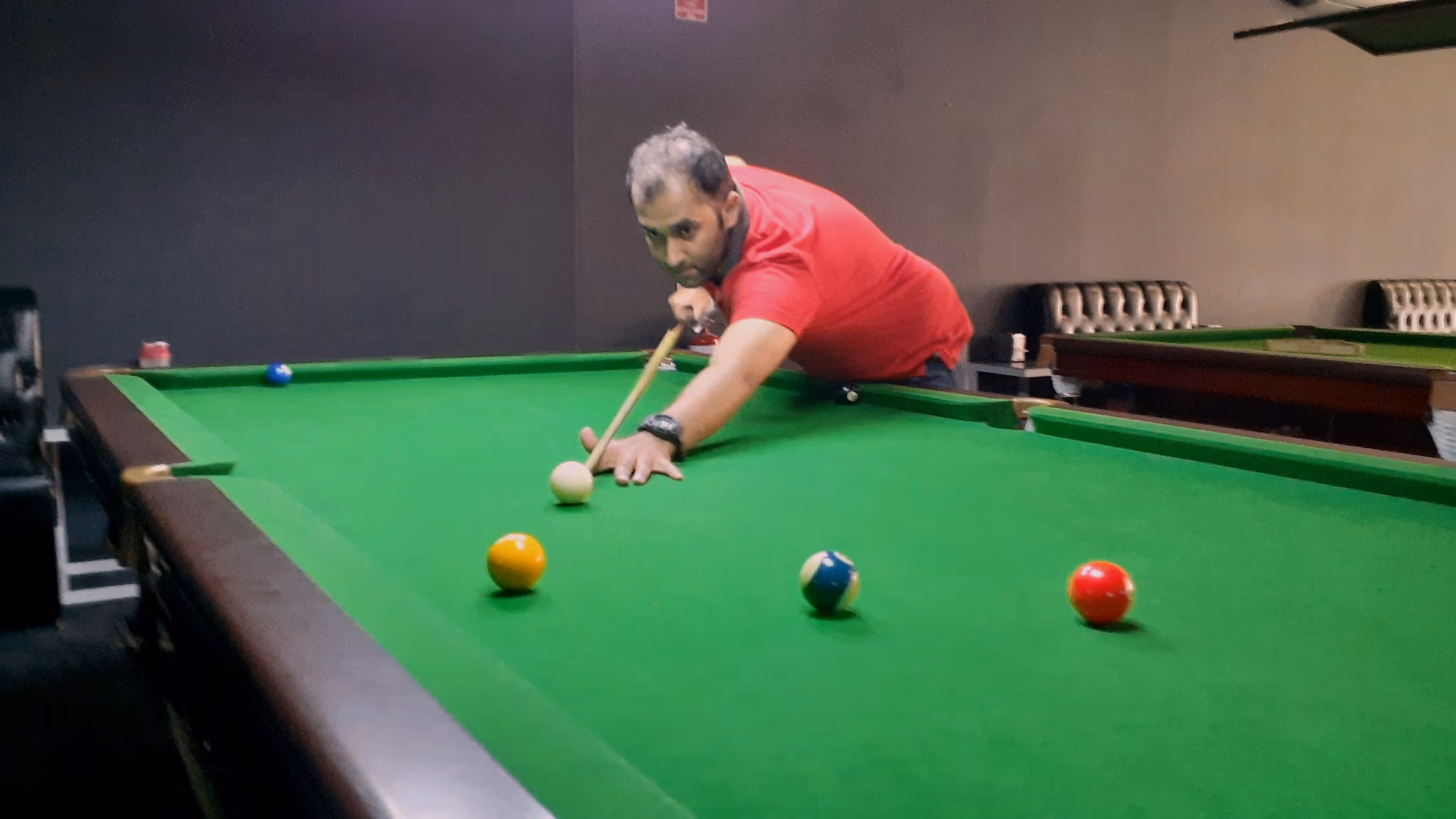 A player sighting along his cue across the green cloth of a full-size snooker table, with the yellow, blue and red balls in front of him.