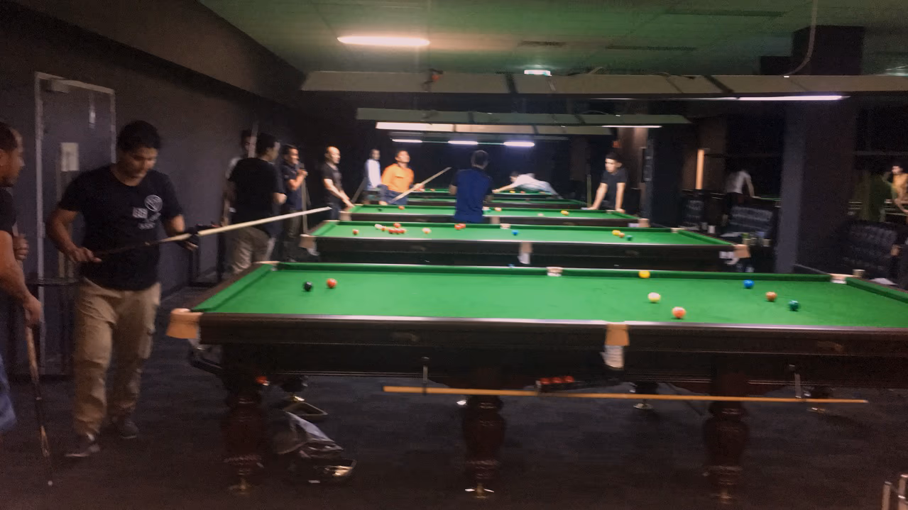 Around a dozen people playing across several full-size snooker tables in a dark room lit by long overhead lamps.